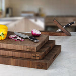 Stack of 3 cutting boards of different thickness with a knife, yellow bell pepper, half a purple onion and onion slices on top. Everything is on a white marble top with grey streaks and a minimalistic knife block is behind the cutting boards.